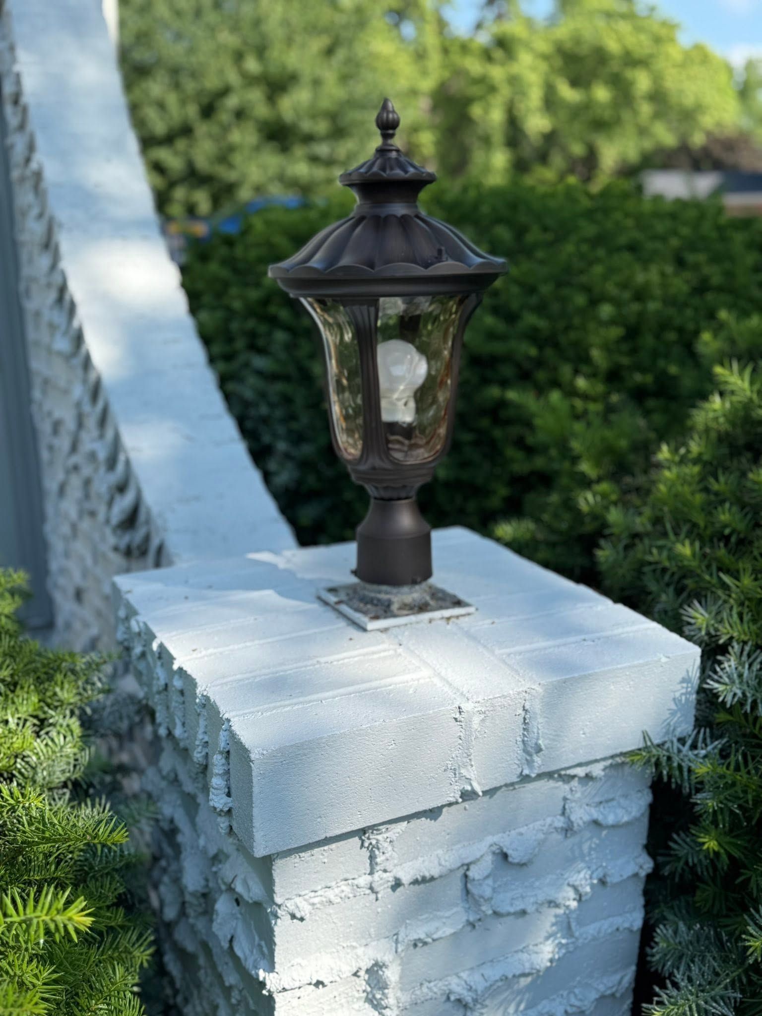 Black outdoor lantern on a white brick pillar beside a stone wall and greenery